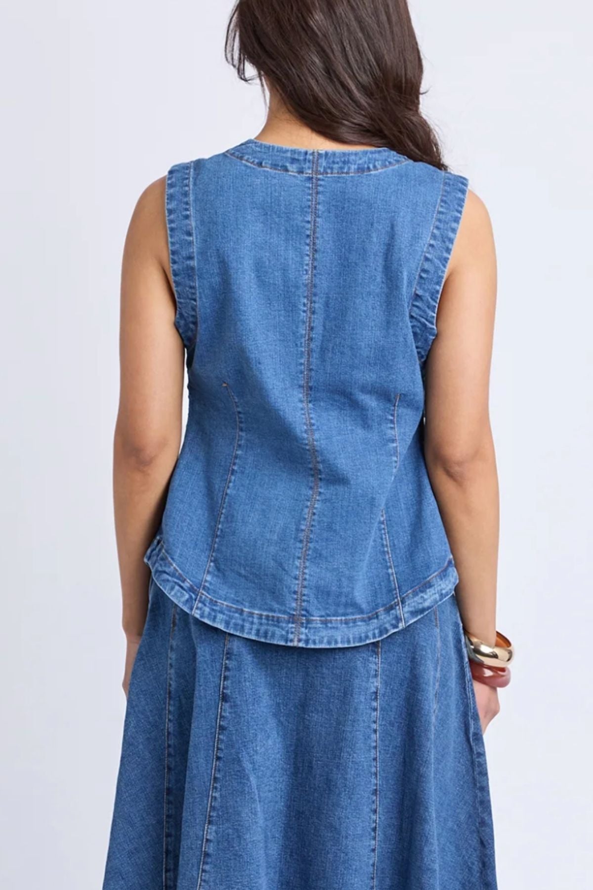 A woman with long dark hair is seen from the back, wearing the sleeveless Foxwood Abbott Vest in blue stretch denim paired with a matching denim skirt, creating a modern look. She accessorizes with a bracelet on her right wrist.