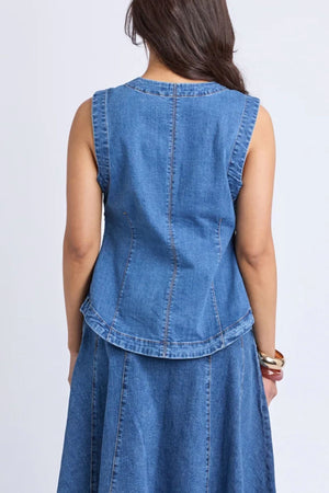 A woman with long dark hair is seen from the back, wearing the sleeveless Foxwood Abbott Vest in blue stretch denim paired with a matching denim skirt, creating a modern look. She accessorizes with a bracelet on her right wrist.