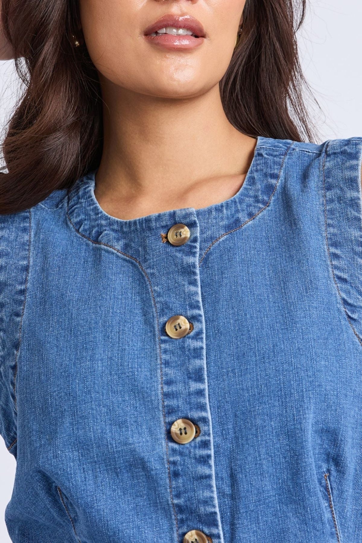 A close-up shows a woman in the Foxwood Abbott Vest, a sleeveless blue stretch denim piece with large tan buttons. Only her lips, neck, and upper torso are visible, emphasizing its modern fit and stylish design.