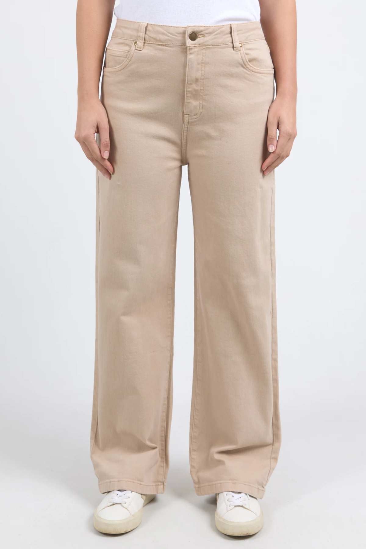 A person wearing beige Foxwood Adira Jeans and a white top is standing against a plain background. The person is also wearing white sneakers. The photograph captures the individual from the shoulders down, highlighting the wide leg fit reminiscent of the Foxwood Adira Jean style.