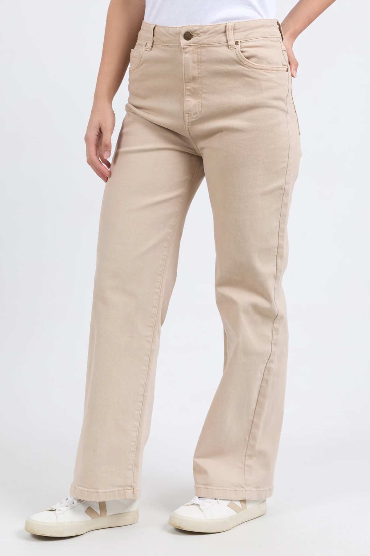A person is standing against a plain background wearing the Foxwood Adira Jean in beige. The high-waisted, wide-leg fit jeans are paired with a white top and white sneakers. With their right hand resting on their hip, they highlight the stylish stretch denim fabric of the Foxwood Adira Jean.