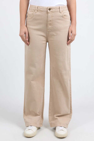 A person wearing beige Foxwood Adira Jeans and a white top is standing against a plain background. The person is also wearing white sneakers. The photograph captures the individual from the shoulders down, highlighting the wide leg fit reminiscent of the Foxwood Adira Jean style.