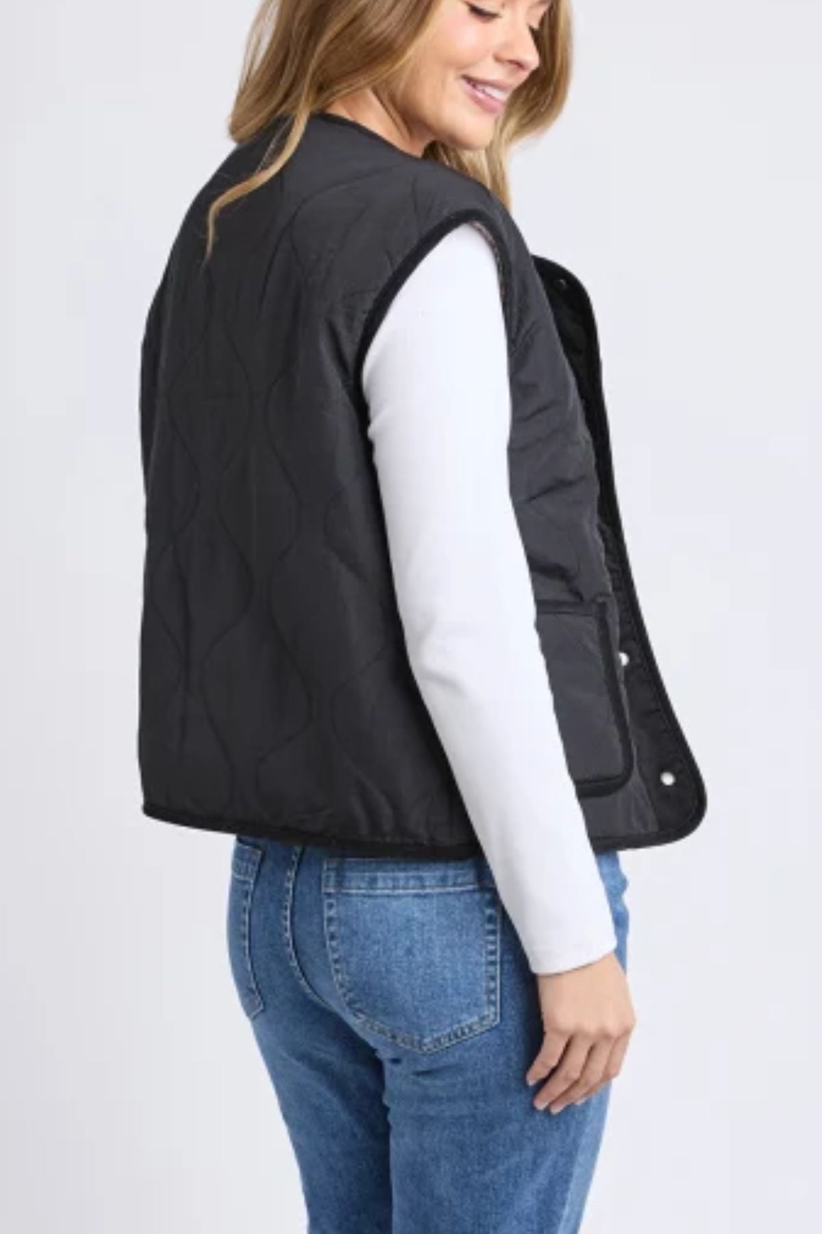 A woman with long, light brown hair is seen from the side and back, wearing the Foxwood Alberta Vest in black with sherpa lining over a white long-sleeve shirt and blue jeans. She is smiling and looking downward.
