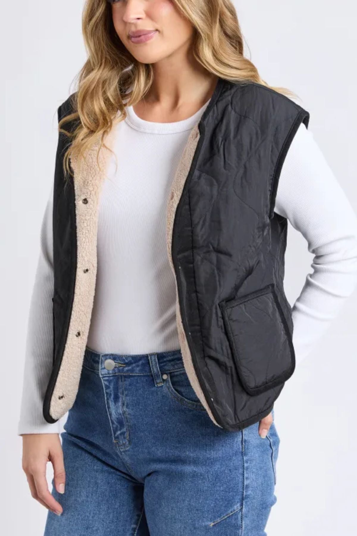A woman wearing a Foxwood Alberta Vest—black, quilted, with sherpa lining and a front pocket—paired with a white long-sleeve top and blue jeans, stands against a plain light background.