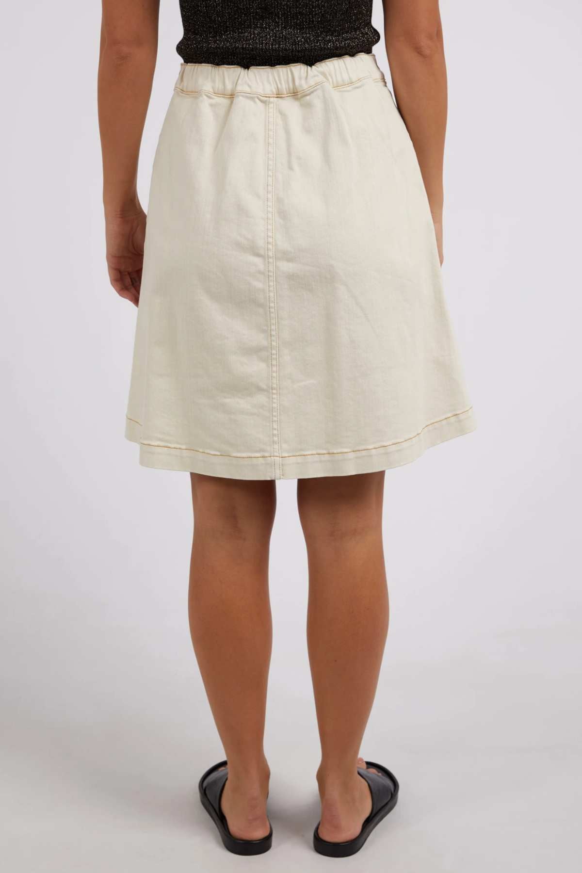 A person wearing the Foxwood Amanda Skirt, a cream-colored A-line with an elastic waistband, paired with a black top and sandals, is shown from the back against a plain light background for a subtle retro vibe.