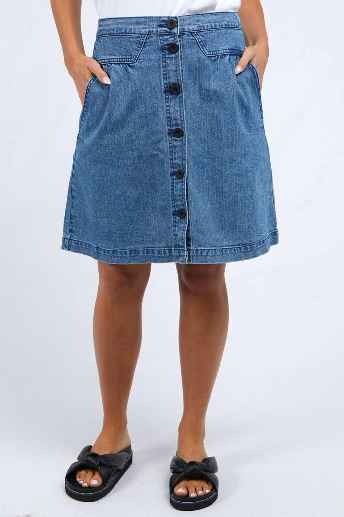 A person is sporting a knee-length Amanda Skirt by Foxwood, featuring front buttons, paired with a white top. They have their hands casually in their pockets and are styled with black open-toed sandals adorned with bow accents, creating a retro look against the simple white backdrop.
