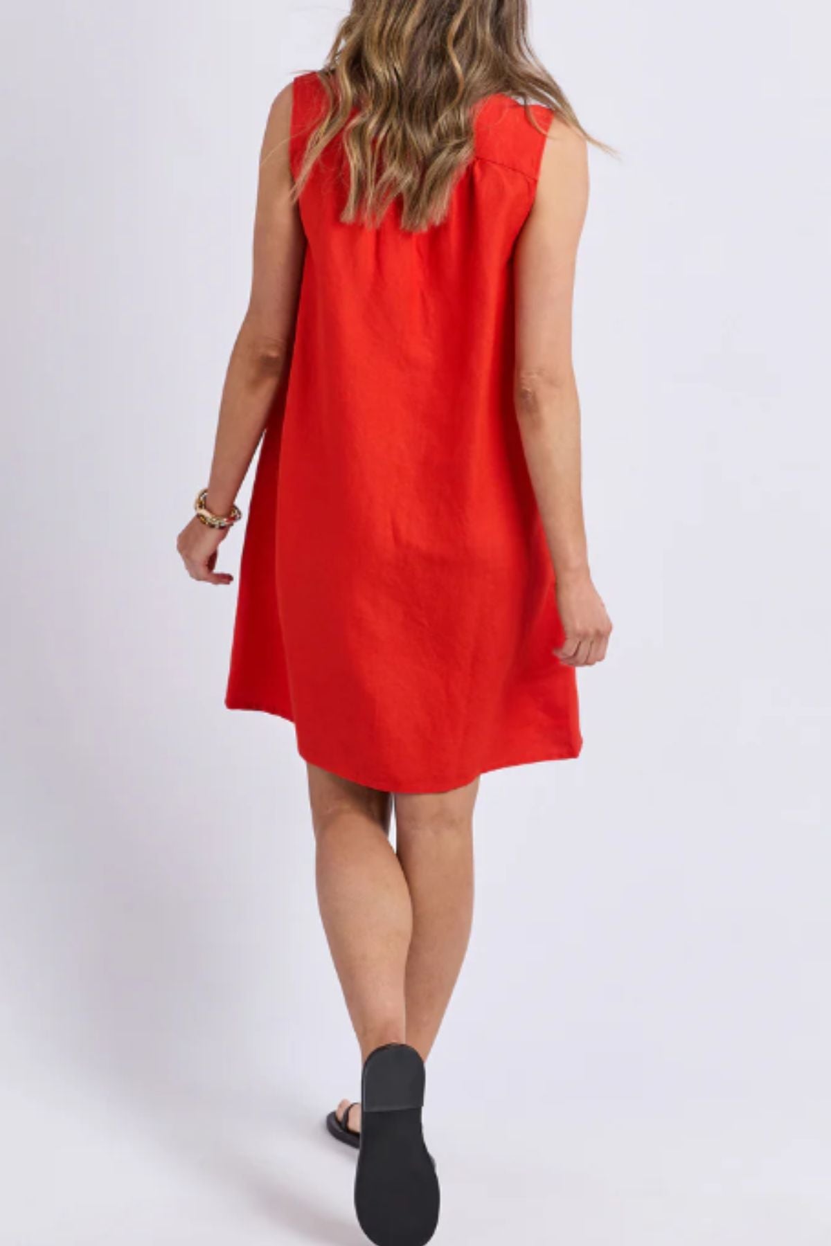 A woman with long wavy hair is shown from behind wearing the sleeveless, loose-fitting Foxwood Anara Dress with a V-neckline and black sandals, walking away against a plain light background.
