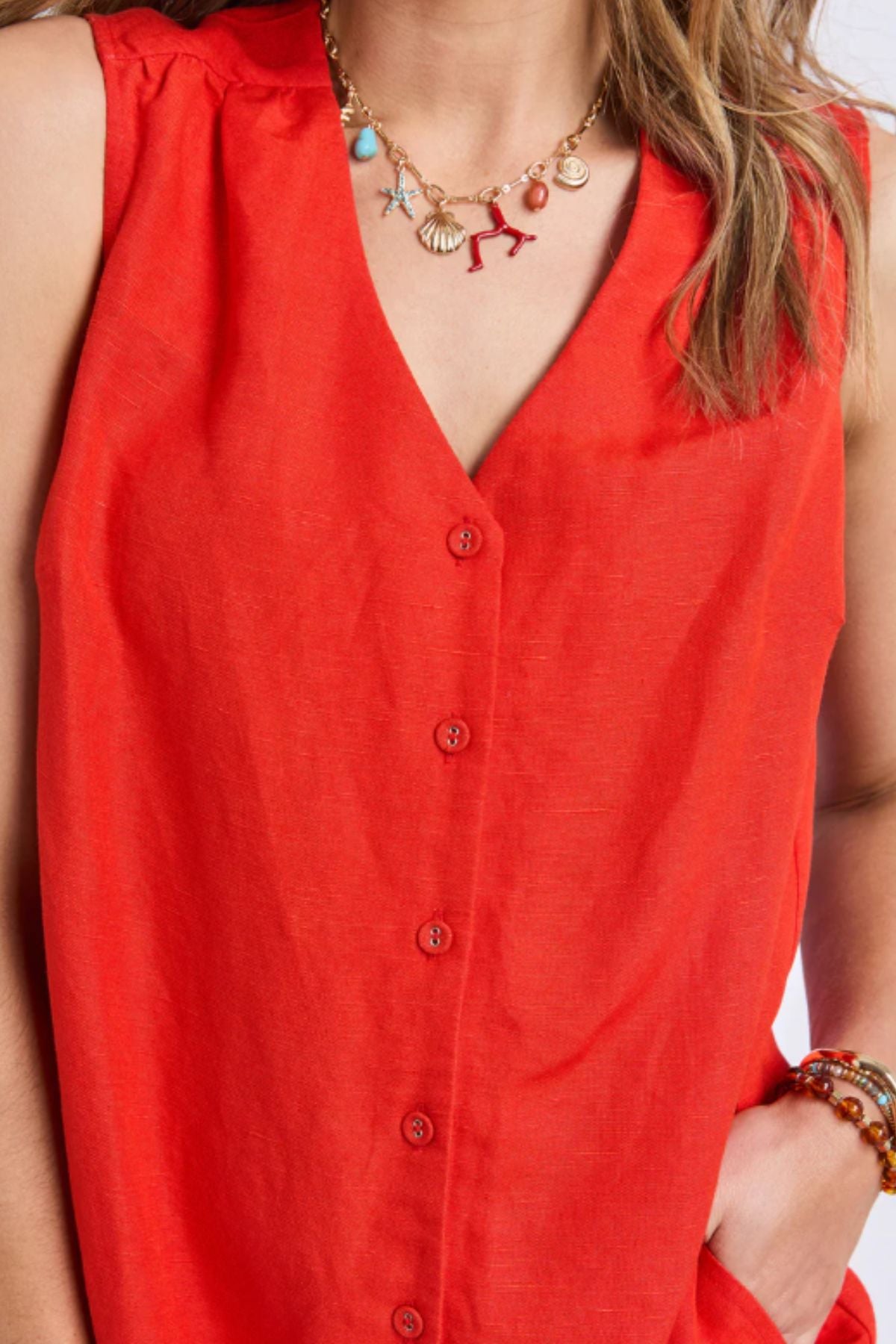 A person wearing the sleeveless, bright red Foxwood Anara Dress with a V-neckline, paired with a beaded bracelet and a gold chain necklace adorned with colorful charms like shells and a starfish.