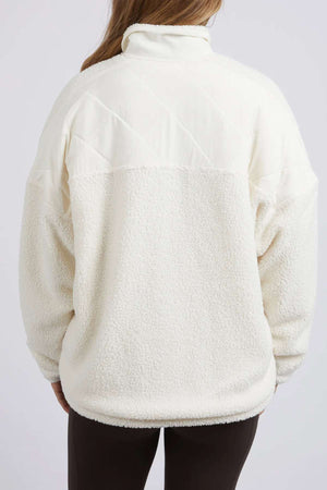 A person with long hair is viewed from the back wearing a Foxwood Arizona Fleece Zip Thru in cream, featuring a high collar and contrasting shearling texture on the bottom half along with smooth quilted fabric on the top. The person pairs it with dark pants.