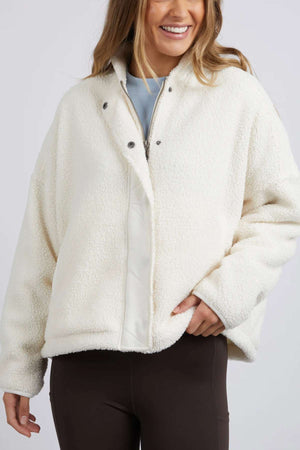 A person with long hair, smiling, is wearing a white Foxwood Arizona Fleece Zip Thru jacket with a snap button front and their right hand in one of the jacket's pockets. They’re also dressed in brown leggings. The background is plain and light-colored.