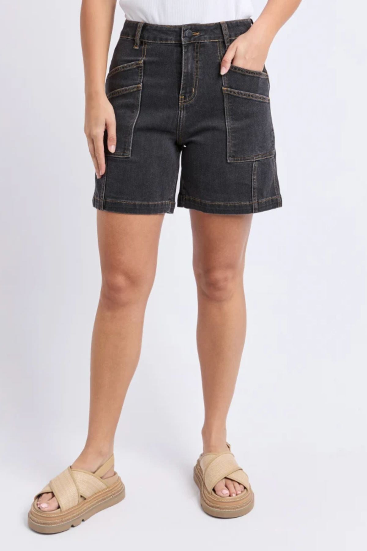 A person models the Foxwood Balmy Short in washed black, featuring front patch pockets, paired with a white top and beige sandals, standing against a light background. Only the body from waist to feet is visible.