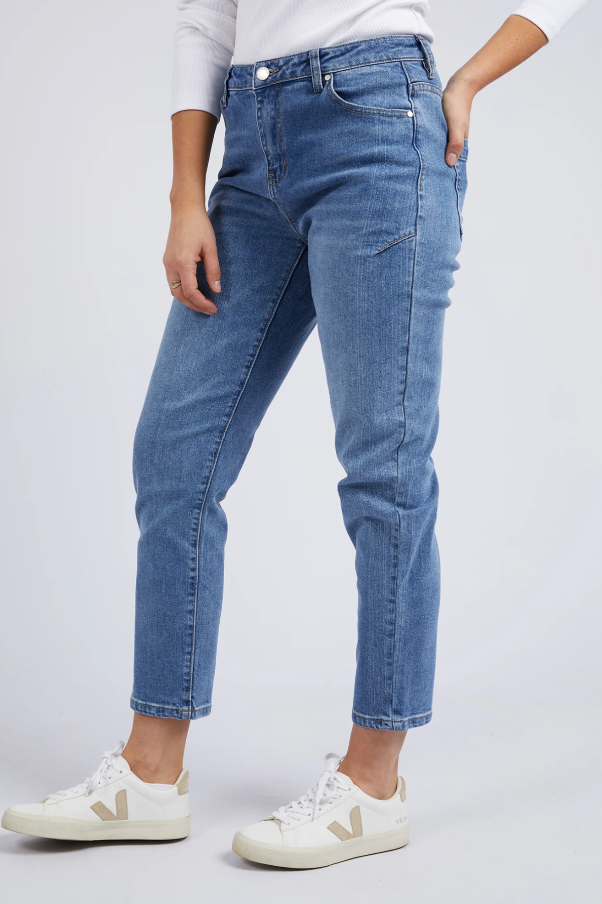 Wearing mid-rise Foxwood Barkly Straight Leg Jean in light blue stretch denim, a white long-sleeve top, and white sneakers with beige accents, a person stands with one hand in their pocket against a plain light background.