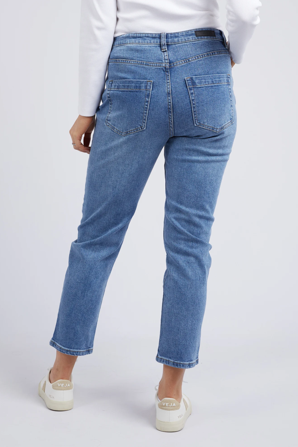 A person wearing the Foxwood Barkly Straight Leg Jean in light blue stretch denim, a white long-sleeve top, and white Veja sneakers is shown from the back against a plain light background.