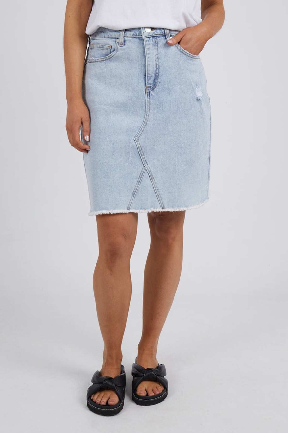 A person stands against a plain background, wearing the Foxwood Belle Skirt in light blue, featuring a frayed hem. The stylish A-line silhouette of the skirt is paired effortlessly with a white top and black sandals, while their casual hand rests in one of the skirt's pockets.