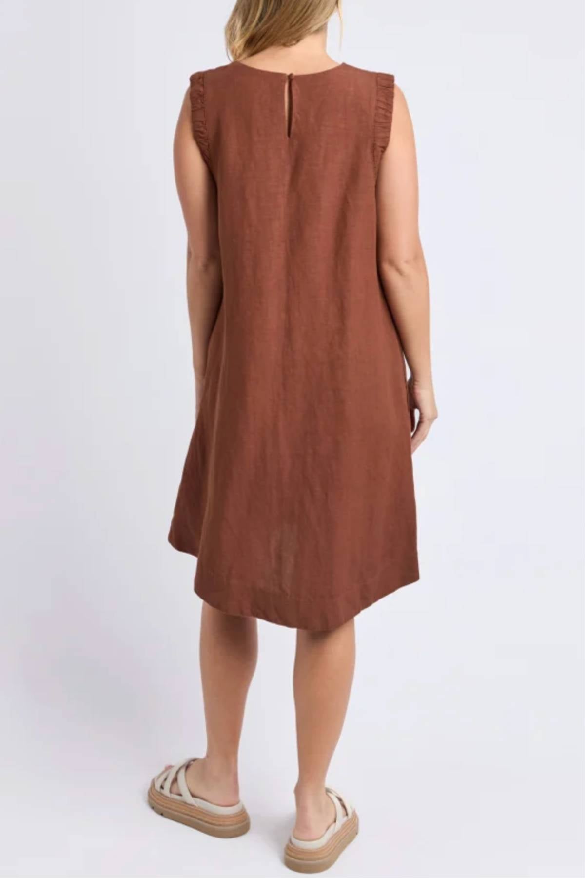 A woman with light skin wears the Foxwood Bermuda Dress—a sleeveless, knee-length brown linen blend dress with a loose fit and ruffled shoulder details. She faces away and pairs it with beige platform sandals.