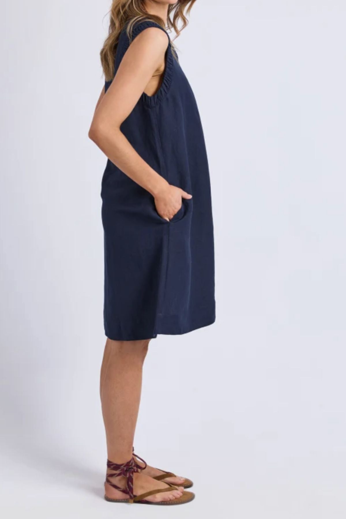 A woman stands in profile against a plain light background, hands in her pockets, wearing the Foxwood Bermuda Dress—a sleeveless, loose-fitting navy linen-blend design—paired with brown flat ankle-strap sandals.