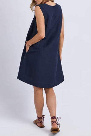 A woman stands facing away, wearing a sleeveless Foxwood Bermuda Dress in navy blue with brown strappy sandals, a bracelet on her wrist, and her left hand in her pocket against a plain light background.