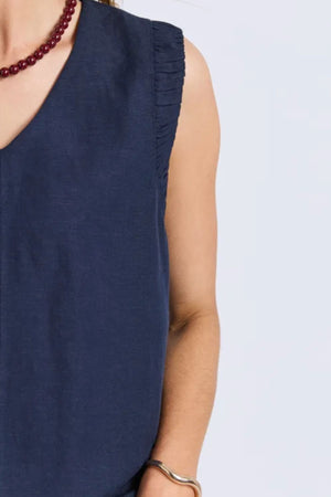 A person wearing the Foxwood Bermuda Dress, a sleeveless navy blue top with frill sleeves and pleated shoulders, paired with a red beaded necklace and gold bracelet. The upper body is shown against a light background.