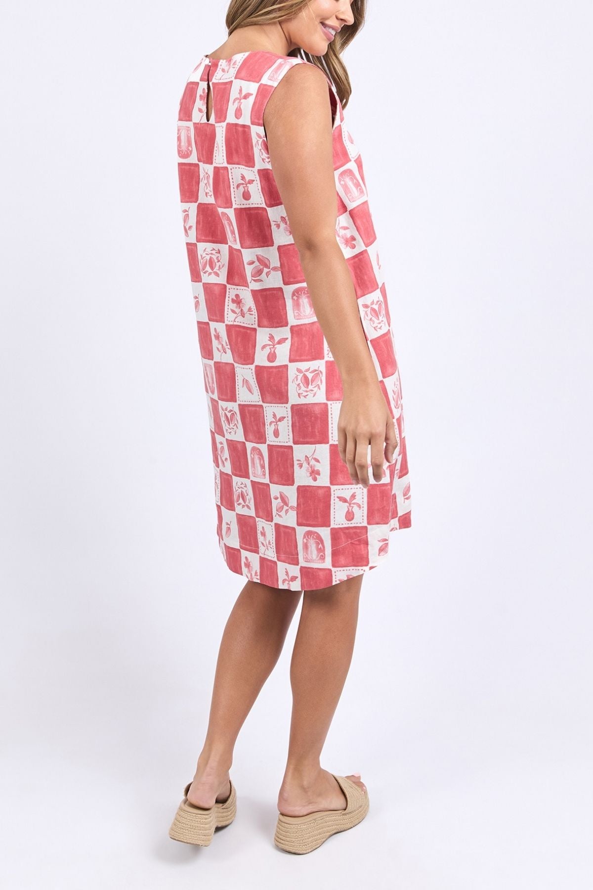 A woman smiles sideways in the Foxwood Biarittz Dress—a sleeveless, knee-length cotton shift in a red and white checkerboard pattern—paired with beige wedge sandals against a plain light background.