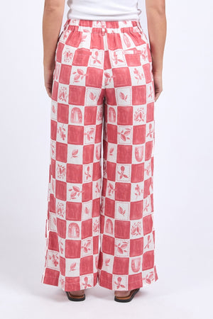 A person wearing the Foxwood Biarittz Pant, featuring red and white checkered fabric with small illustrations, is seen from the back. The pants are made from a linen-viscose blend and include an elastic waistband and side pockets.