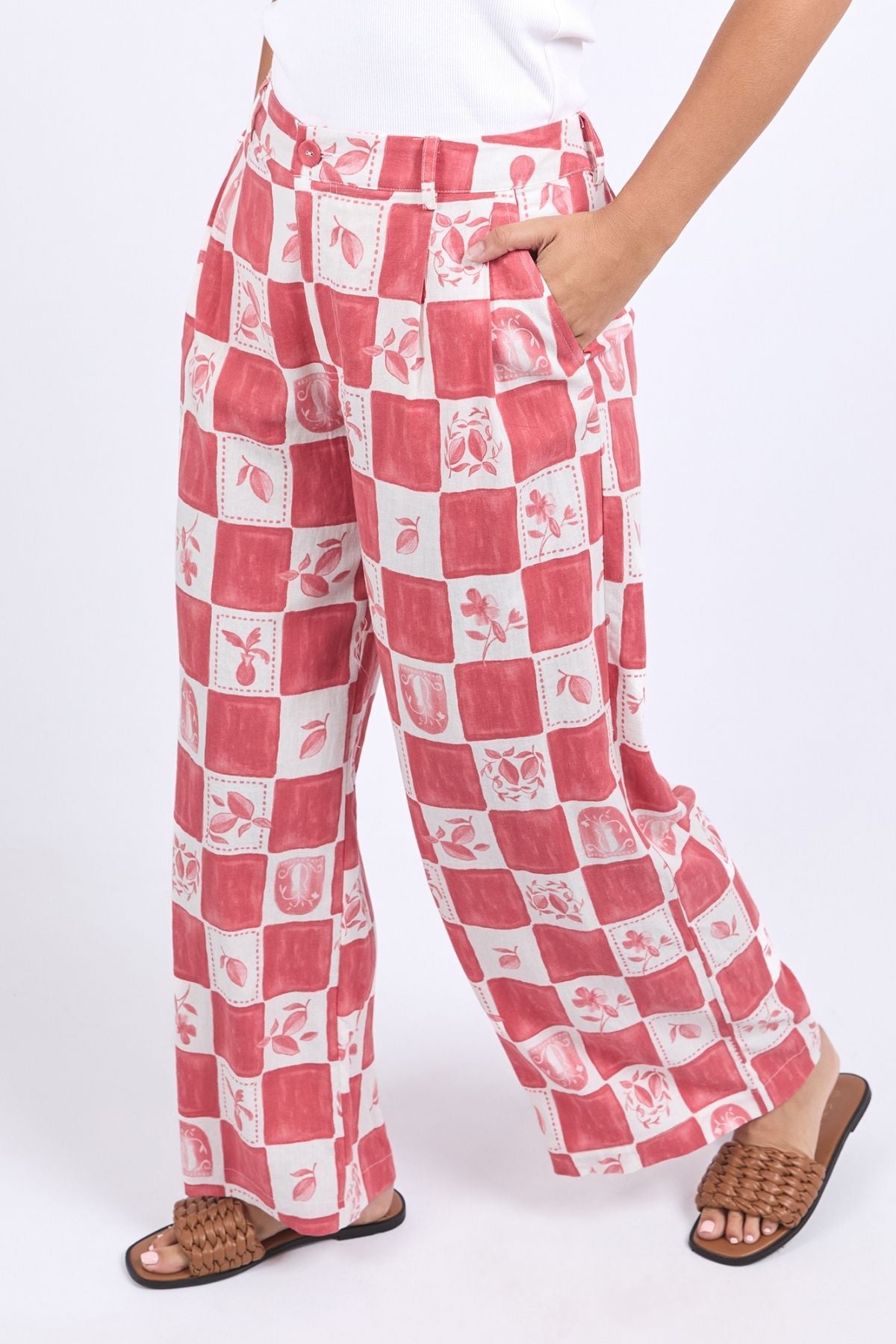 Wearing loose-fitting Foxwood Biarittz Pant with a red and white checkered floral pattern, one hand in the pocket, paired with a white top and brown woven sandals. Made from a soft linen-viscose blend for comfort all day.