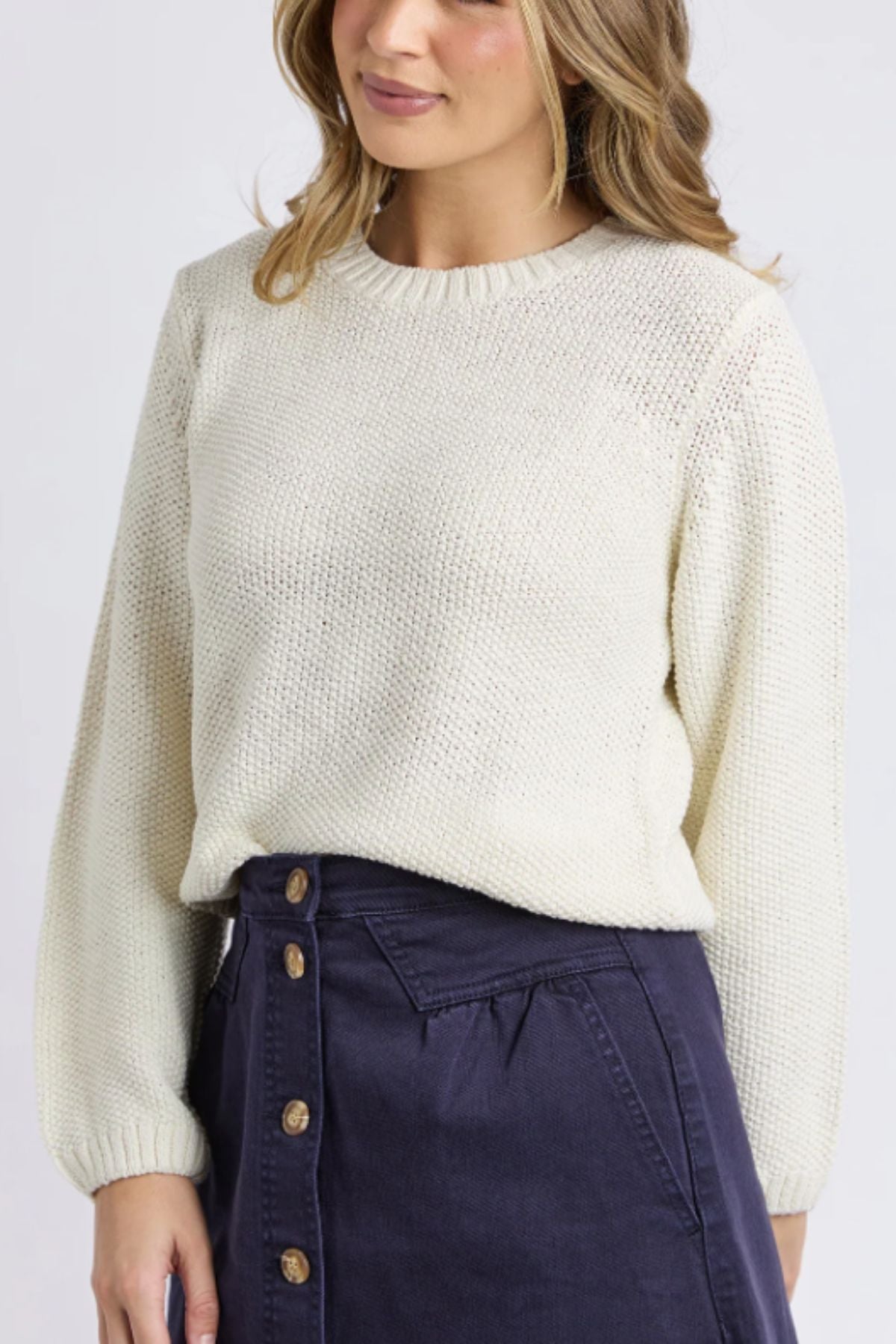A woman wearing the Foxwood Blair Long Sleeve Knit sweater in cream and a navy blue button-front skirt, standing against a light background, shown from mid-thigh up.