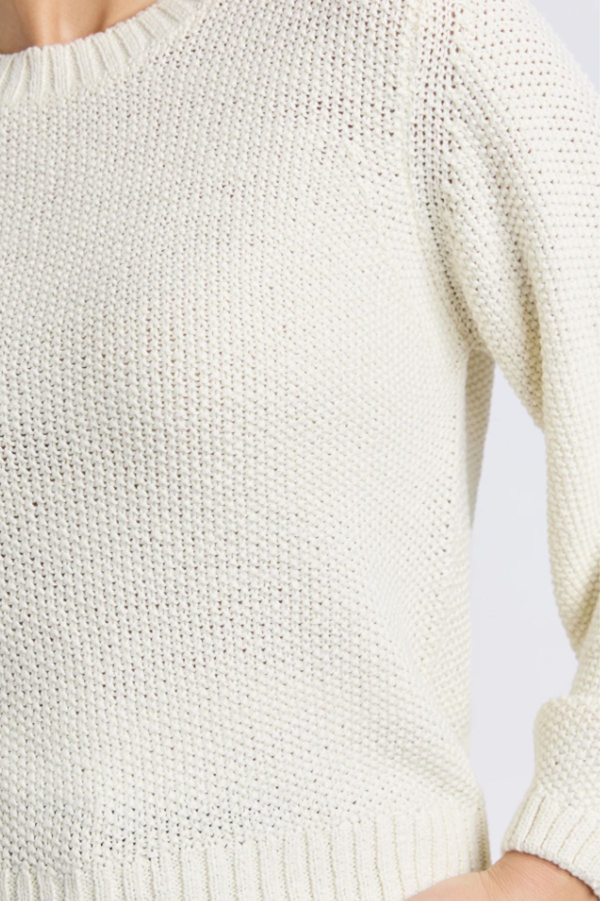 Close-up of a person wearing the Foxwood Blair Long Sleeve Knit, a cream-colored, relaxed-fit sweater with a round neckline and ribbed hem and cuffs. The image highlights the upper torso, shoulder, and part of the left arm. Made from 100% cotton.