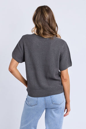 A woman with wavy brown hair, seen from behind, wears the Foxwood Blair Short Sleeve Knit in textured dark gray 100% cotton with light blue jeans, standing against a plain light background.