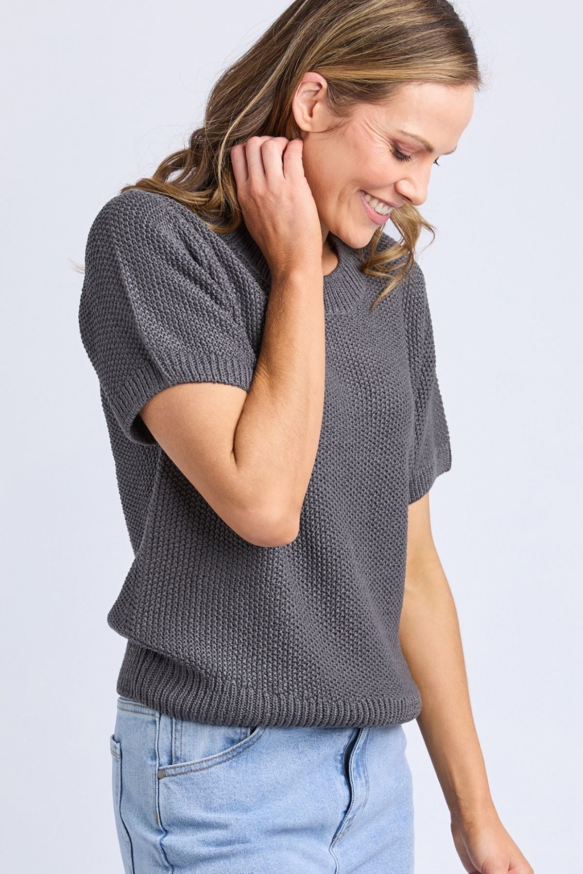 A woman with wavy brown hair smiles as she looks down and touches her neck, wearing a relaxed fit Foxwood Blair Short Sleeve Knit sweater made from 100% cotton, paired with light blue jeans against a light background.