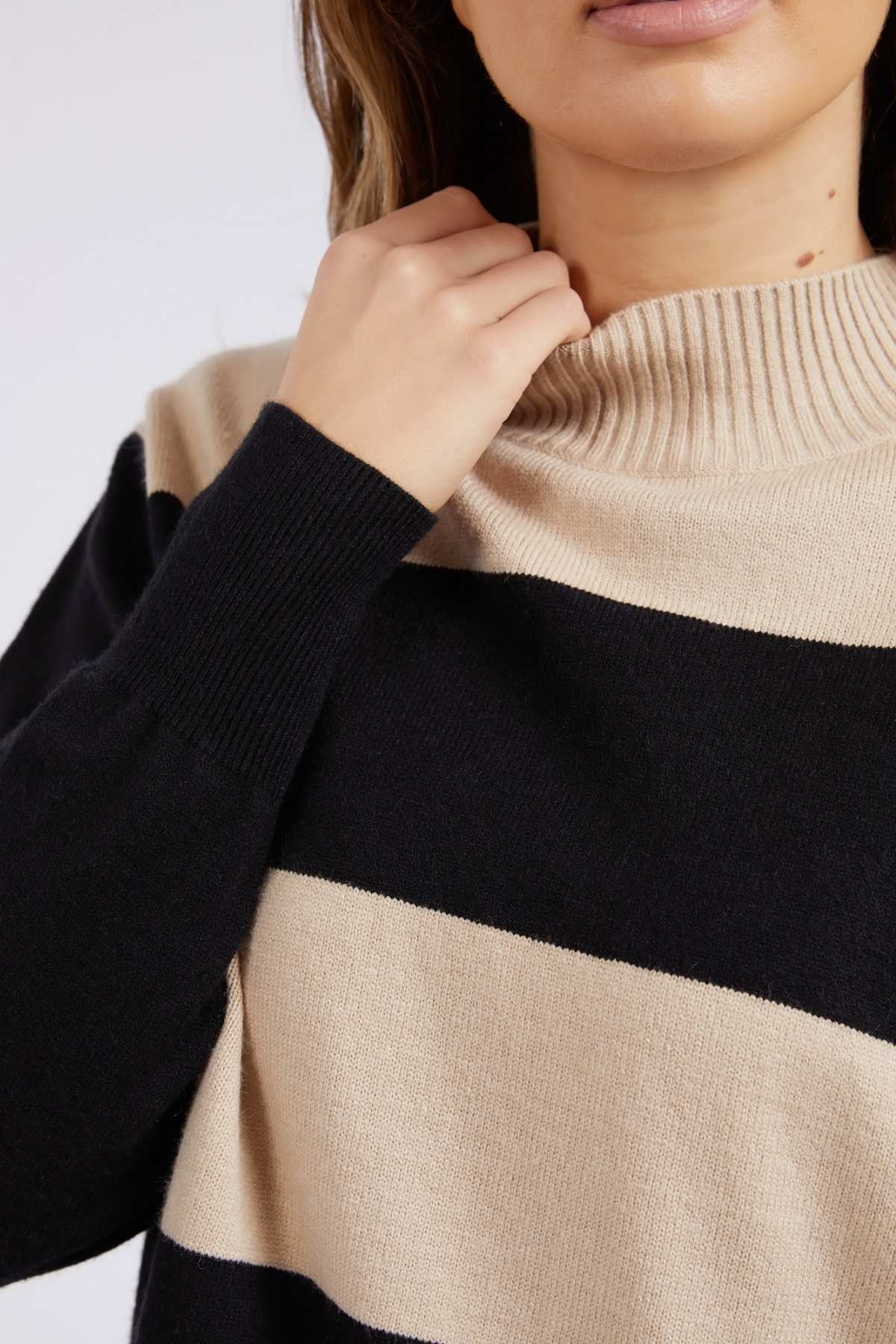 Wearing the Foxwood Canterbury Knit with wide black and beige stripes, their hand near the ribbed collar, the person's look highlights the luxurious wool/cashmere texture against a plain light background.