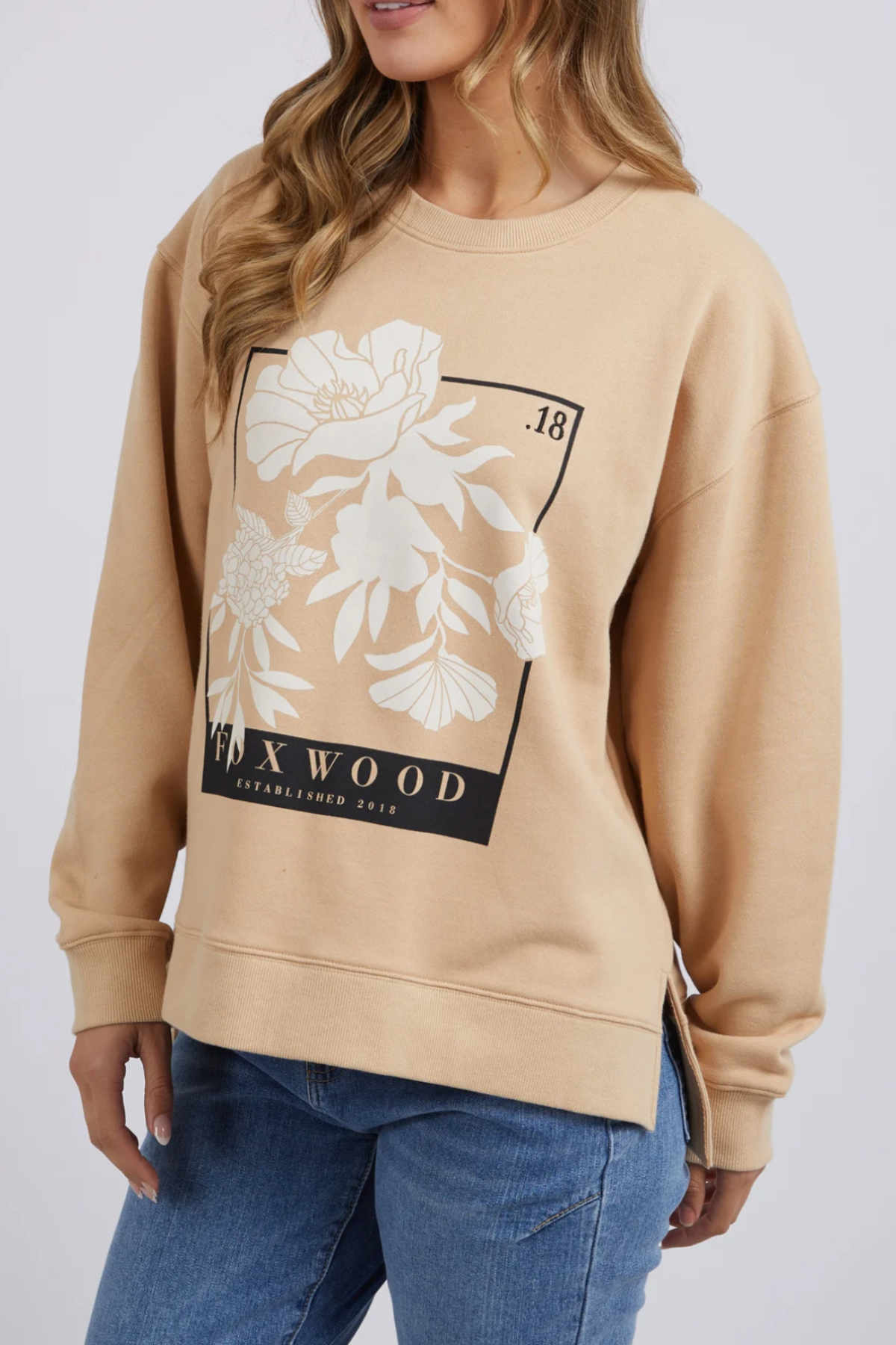 A woman clad in the Foxwood Carnation Crew sweatshirt, featuring a beige color with a floral design and "FOXWOOD" prominently displayed beneath it. The 100% cotton sweatshirt boasts a relaxed fit and includes a side slit, perfectly matched with her blue jeans. Her long, wavy light brown hair cascades as she poses slightly angled to the side.