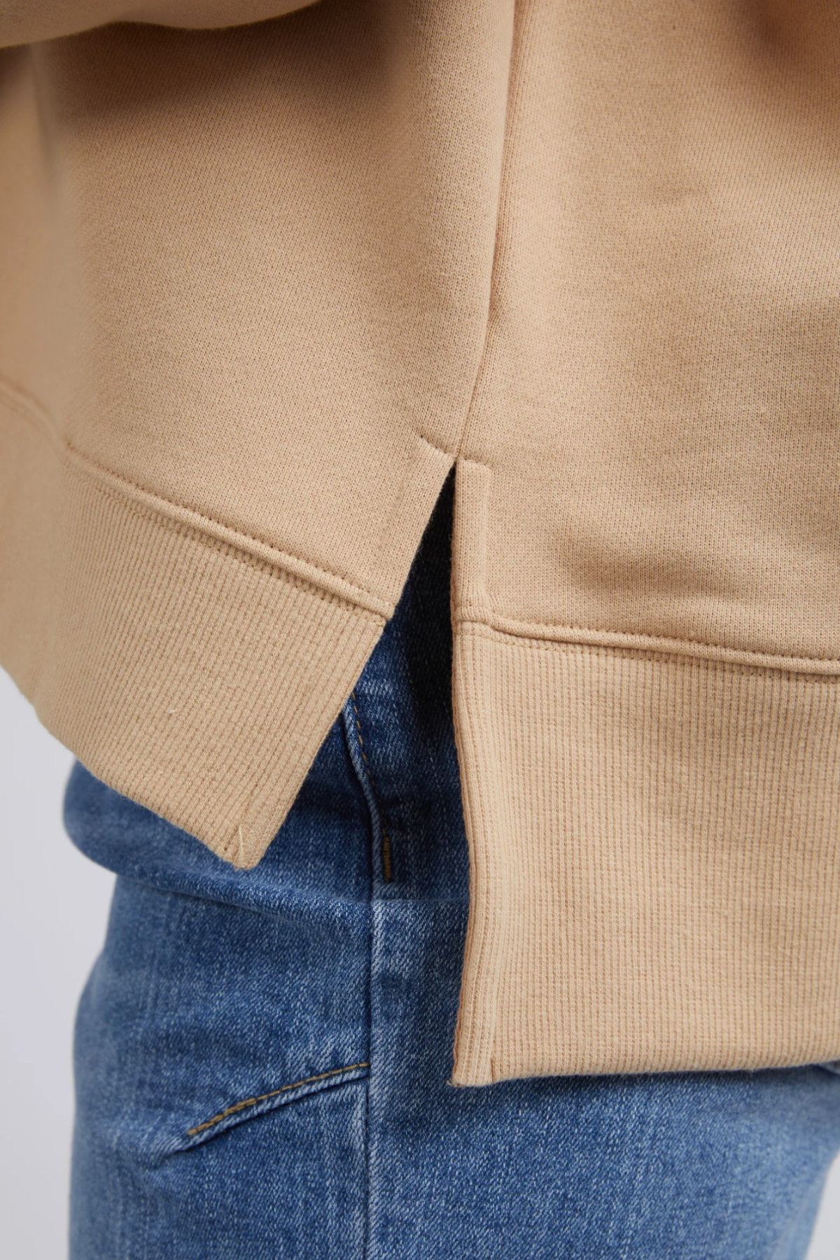 Close-up of the side of a person wearing a beige Foxwood Carnation Crew and blue jeans. The crew features a small side slit at the hem, revealing a clean, finished seam. The texture of the 100% cotton fabric is visible, showcasing its soft, cozy material against the casual blue denim.