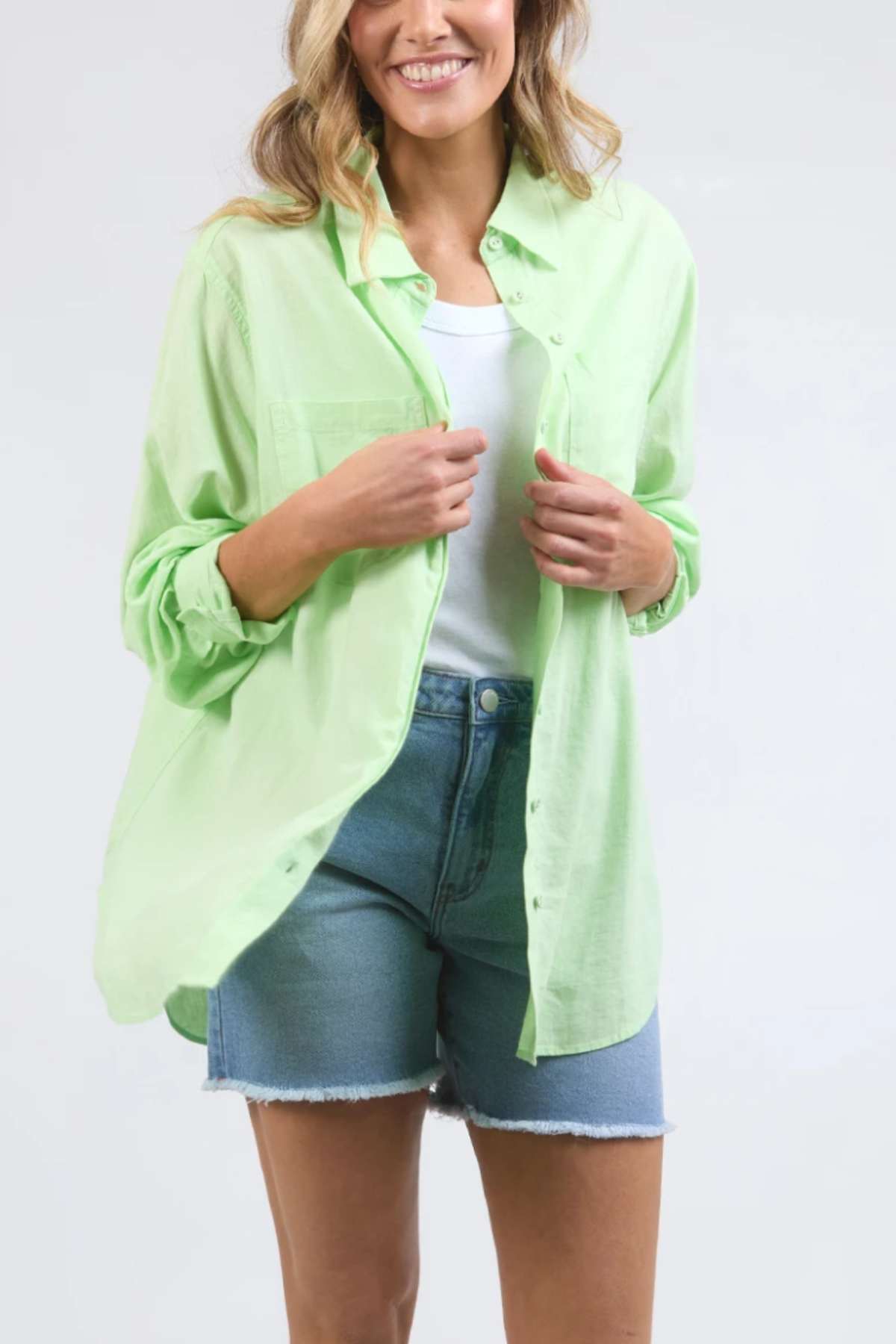 A woman is wearing a light green Foxwood Carrie Shirt over a white top and denim shorts. Crafted from lightweight fabric, the shirt provides a casual, relaxed fit. She has wavy hair that is partially visible. Smiling, she adjusts her Foxwood Carrie Shirt against a plain, light-colored background.