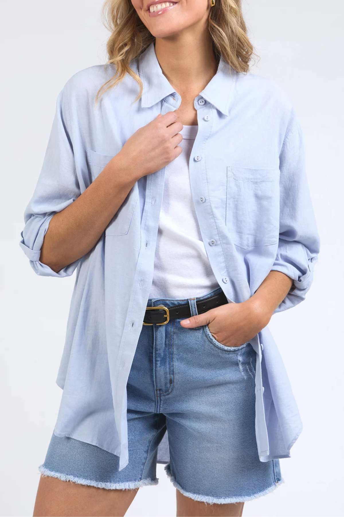 A person wearing a light blue Foxwood Carrie Shirt over a white top is shown from the shoulders to mid-thigh. They have one hand in the pocket of their blue denim shorts, which feature a frayed hem, and the other hand is pulling on the collar of the relaxed fit shirt. The individual has wavy blonde hair.