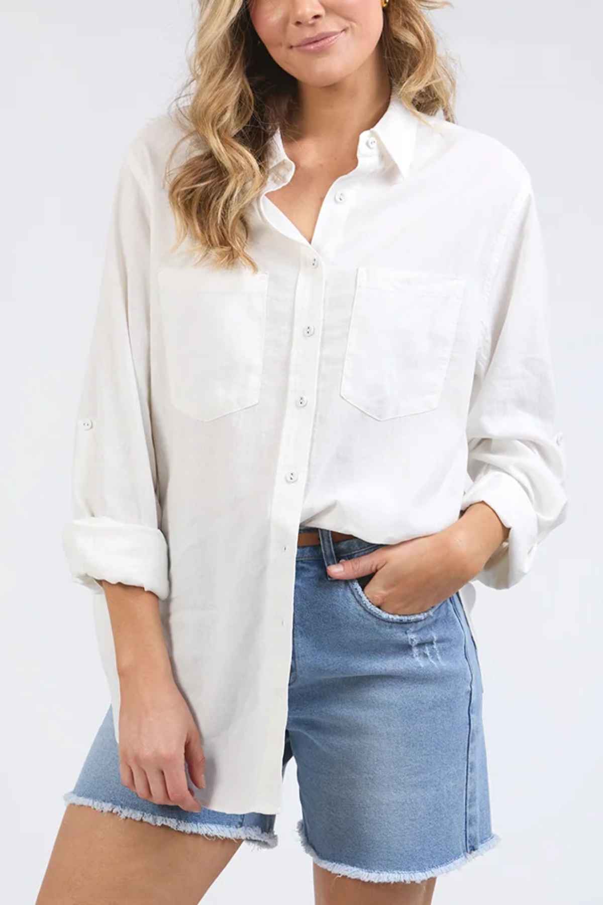 A person sporting the relaxed fit of a Foxwood Carrie Shirt with rolled-up sleeves, teamed with blue denim shorts. The crisp linen fabric beautifully complements the plain white backdrop.