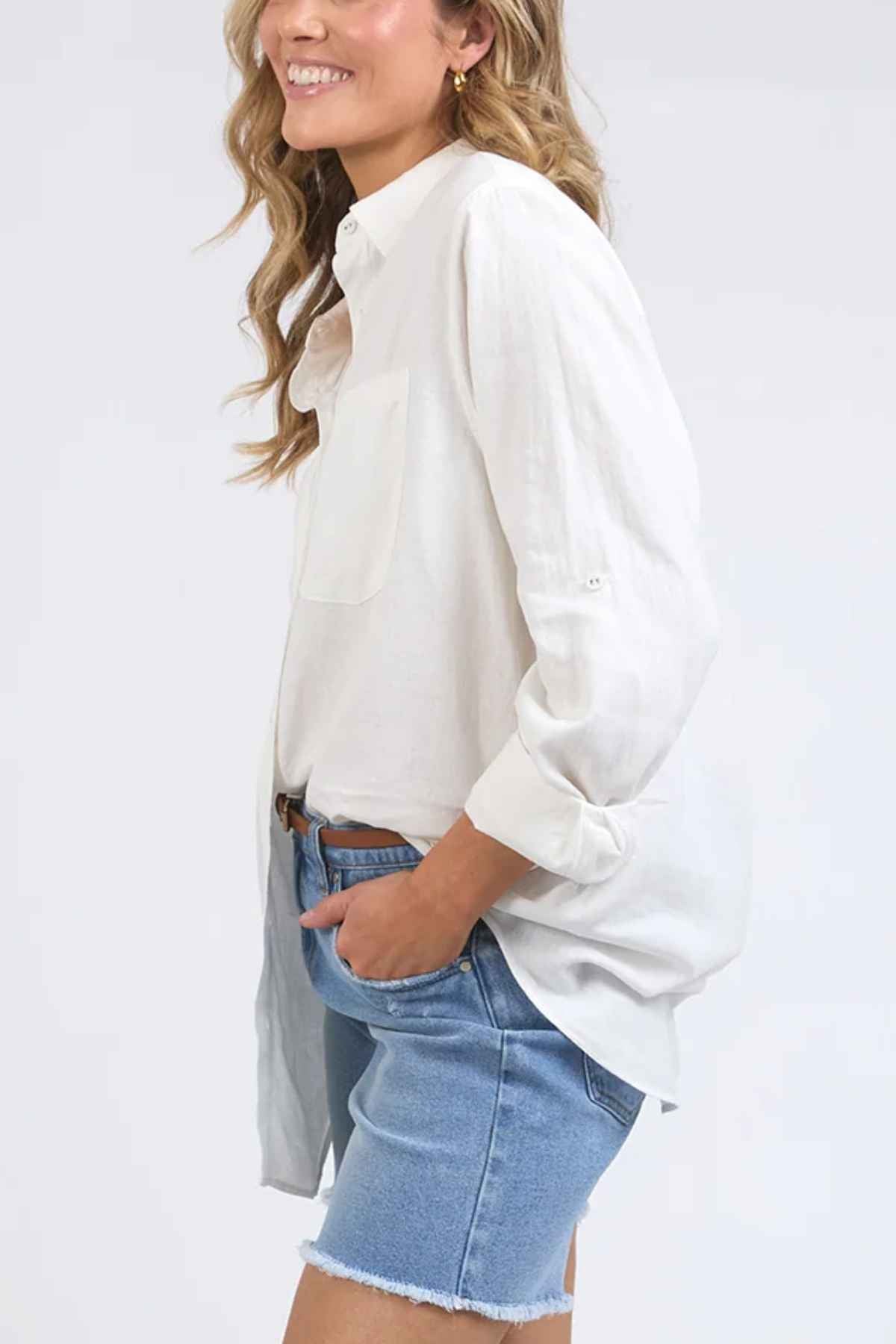 A person in a relaxed fit Foxwood Carrie Shirt and denim shorts, with one hand in their pocket, smiles against a light background.