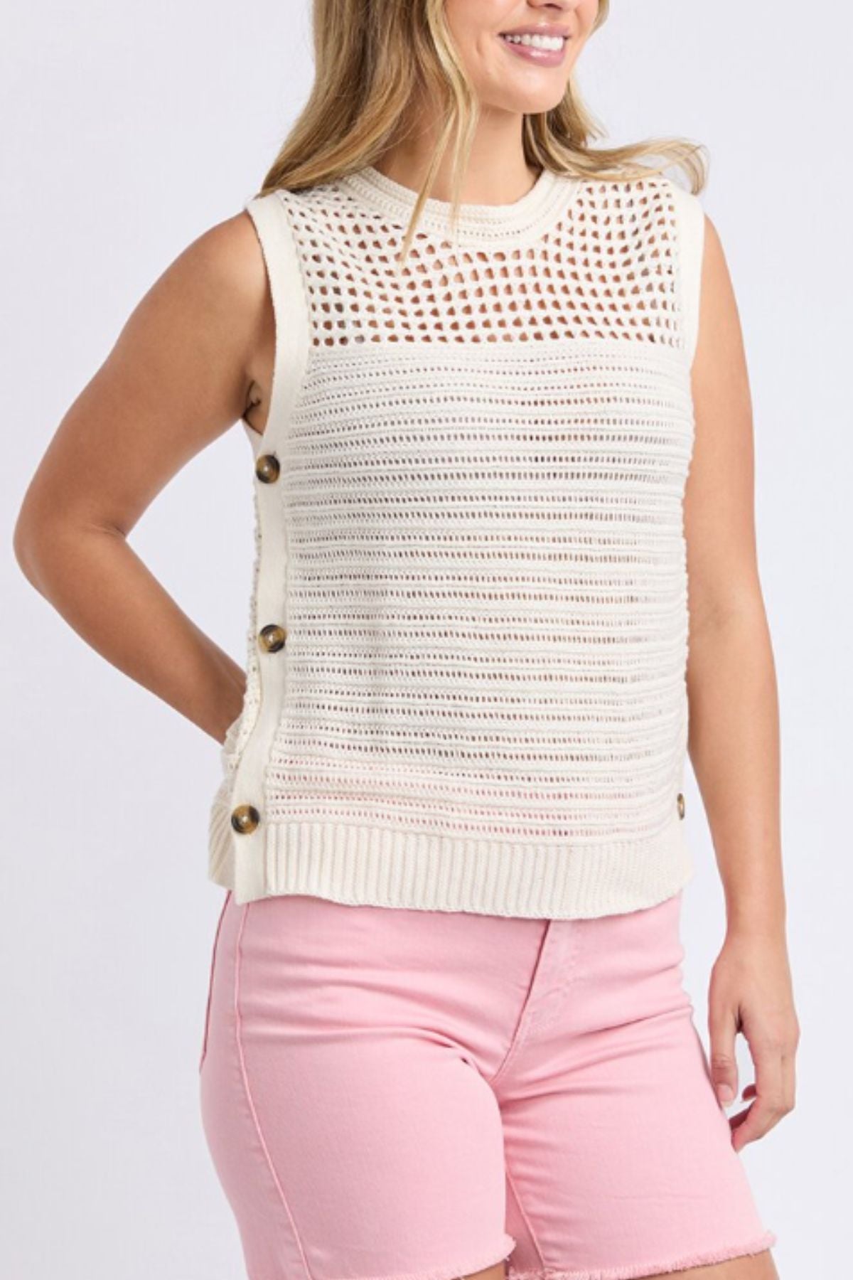 A woman wears the Foxwood Chilli Knit Top, made from 100% cotton stretch knit with large side buttons and a mix of open and closed patterns, paired with light pink shorts. She smiles while standing against a plain background.