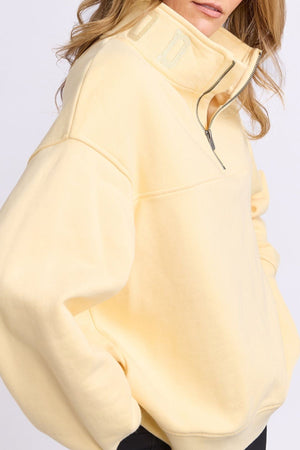 Someone in a loose, pale yellow Foxwood College Zip Crew with a high collar and zip stands with one hand in their pocket. Their wavy hair is down, and their face is partially visible.