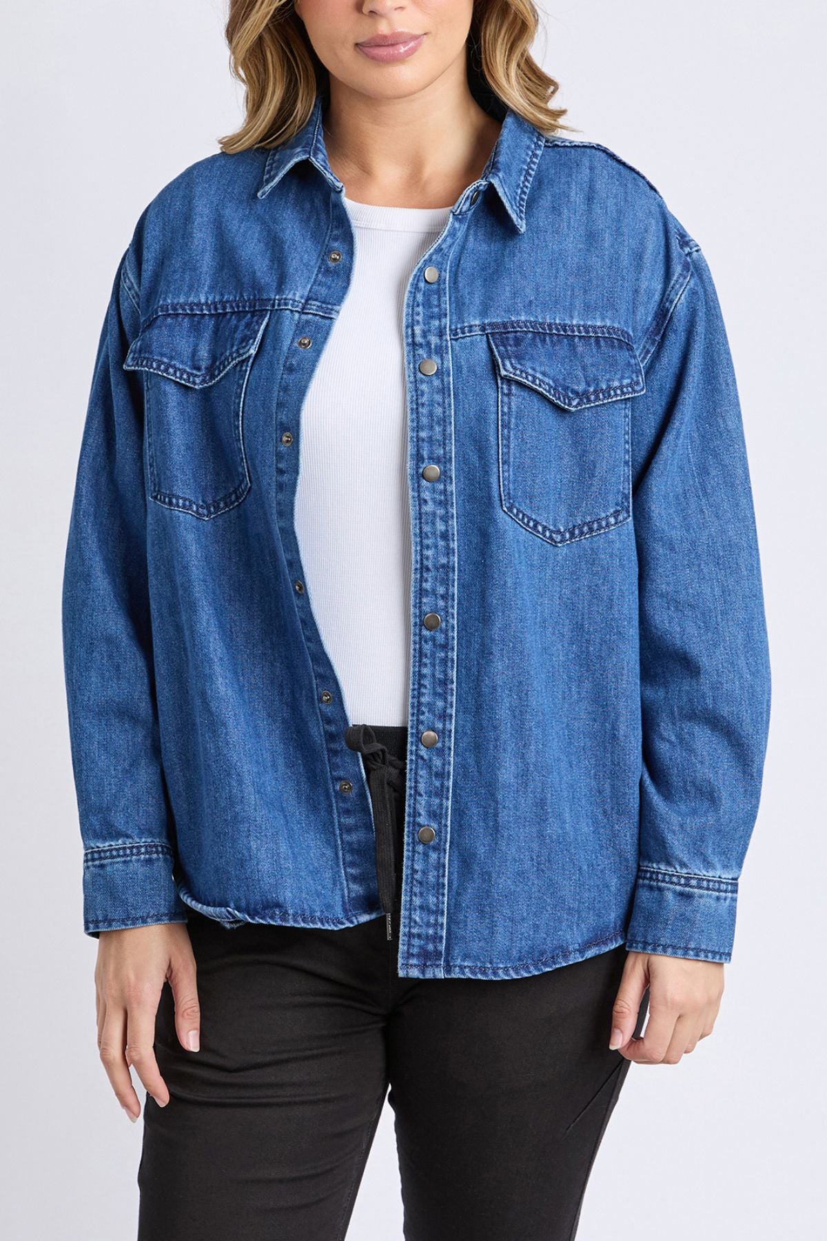 A person wears a relaxed fit, blue denim Foxwood Daisy Overshirt over a white top and black pants, standing against a plain light background. The cotton shirt includes two chest pockets with flaps.