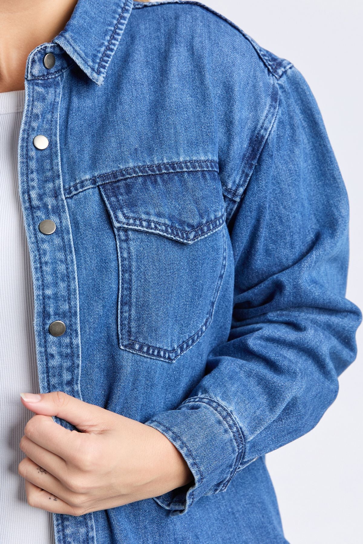 Close-up of a person in a relaxed fit Foxwood Daisy Overshirt—blue denim with metal buttons and front pocket, layered over a cotton shirt. One hand holds the overshirt near the chest.
