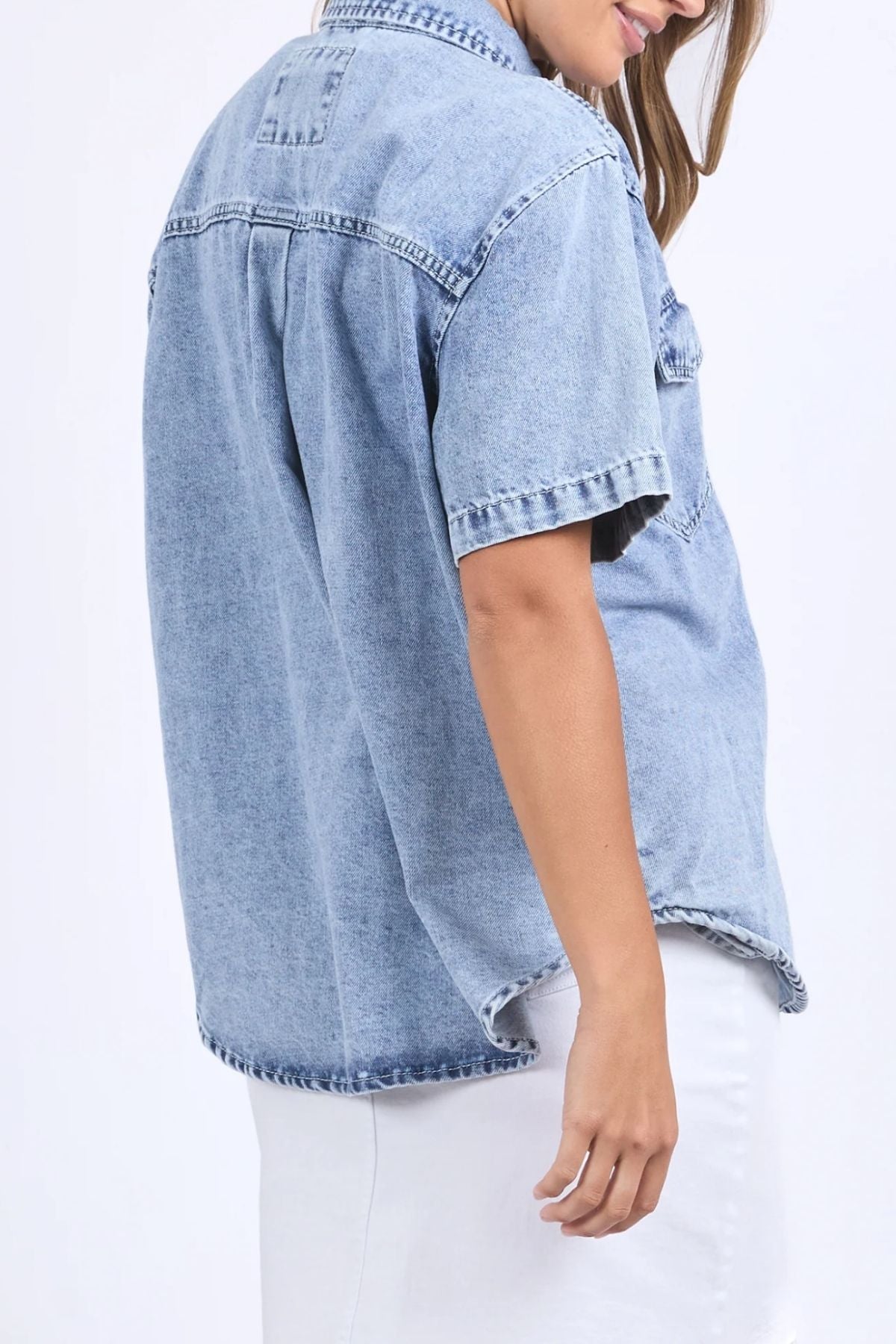A person wearing a relaxed fit Foxwood Daisy Short Sleeve Shirt in light blue denim with a white skirt, standing sideways, head slightly turned, hair partially covering their face.