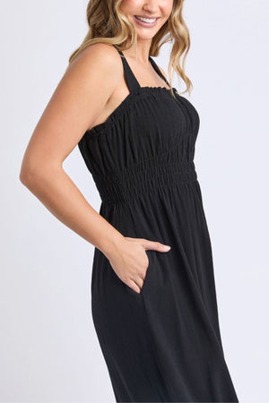 A woman in the Foxwood Dandelion Dress—a sleeveless black linen blend with shirred waist and bust—stands sideways with a hand in her pocket, smiling slightly. The light, plain background highlights this perfect summer dress.