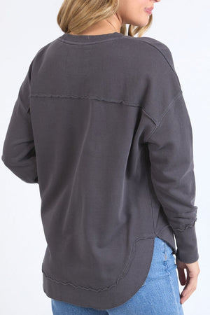 A woman with long blonde hair, wearing a dark gray Foxwood Delilah Crew jumper made from 100% cotton and light blue jeans, is shown from the back against a plain light background.