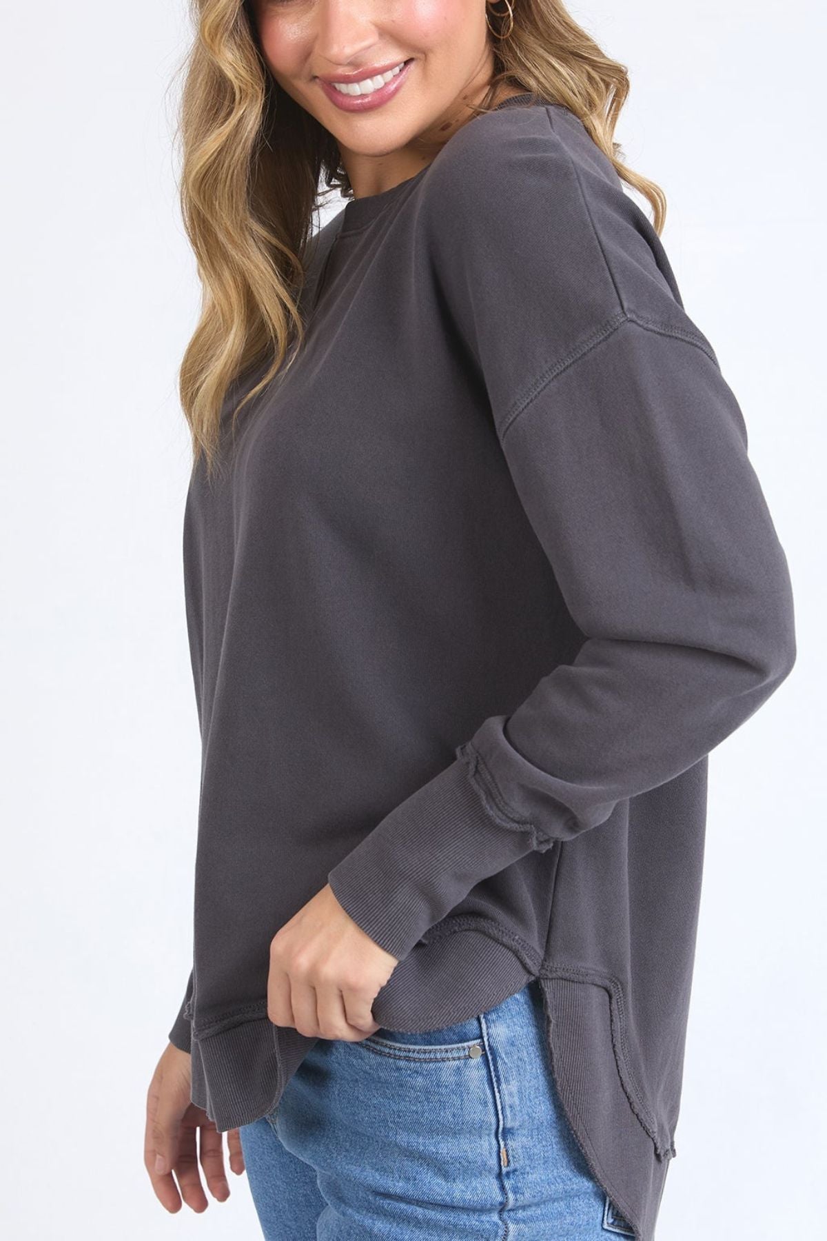 A woman with long, wavy hair, wearing a loose-fitting dark gray Foxwood Delilah Crew and light blue jeans, smiles while standing sideways against a plain white background.