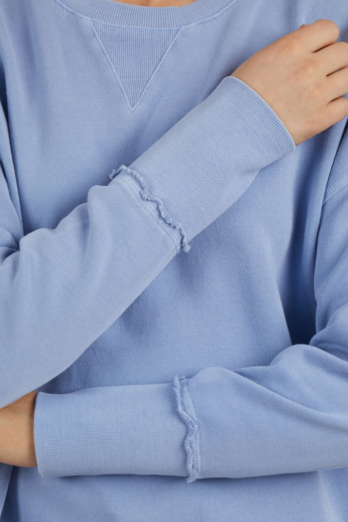 A person in a Foxwood Delilah Crew, a light blue cotton jumper with visible stitching on the sleeves, stands with arms crossed and one hand on the opposite shoulder. The neutral, out-of-focus background emphasizes the jumper's relaxed style.