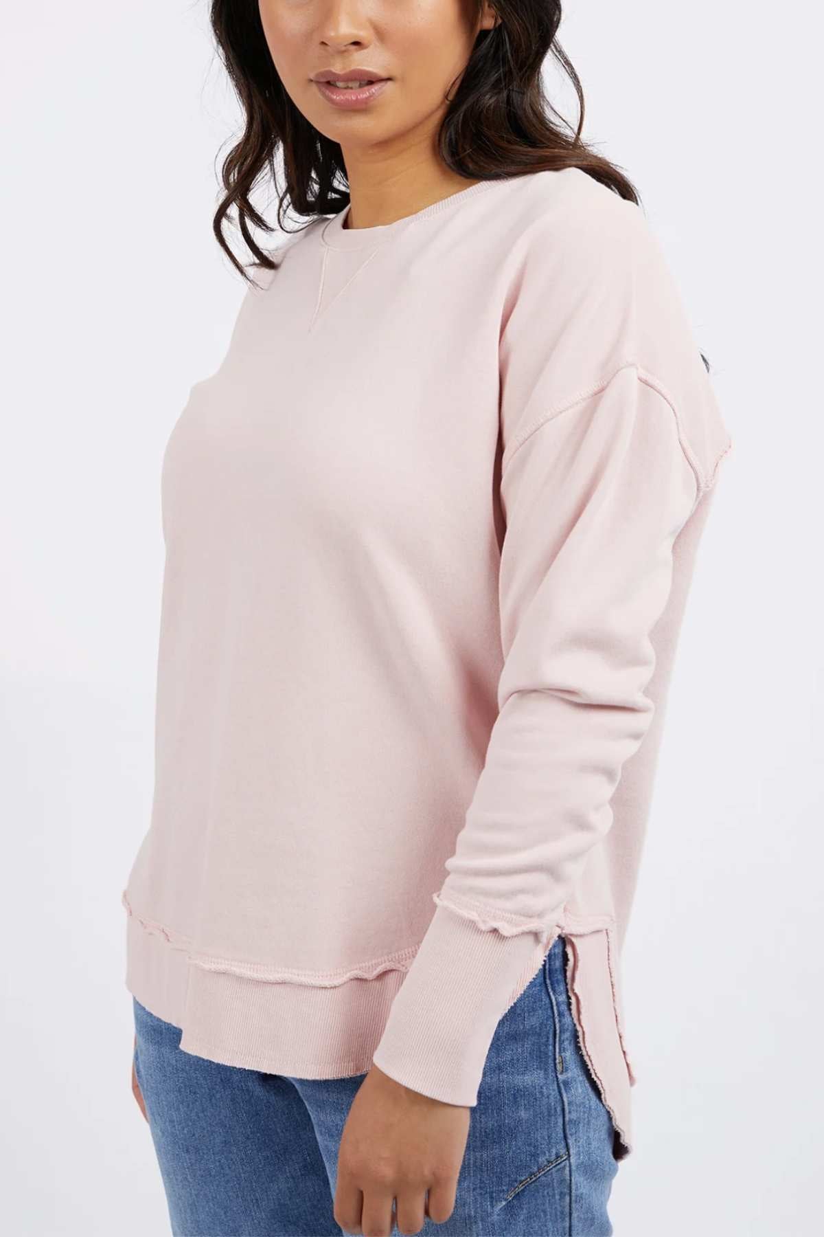 A woman wearing the Foxwood Delilah Crew, a light pink cotton jumper with raw edges, and blue jeans stands against a plain white background.