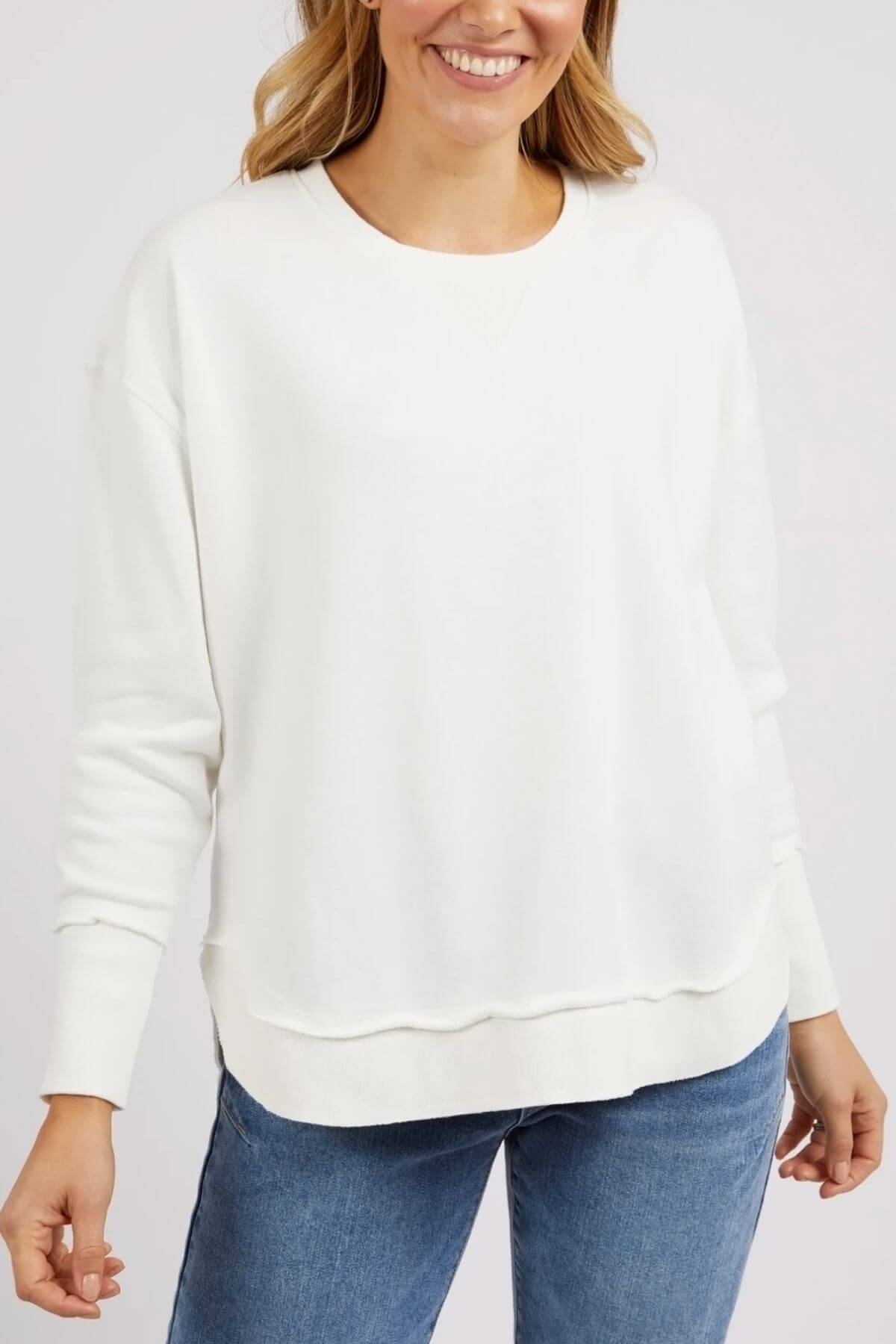 A woman wears a loose-fitting, long-sleeve Foxwood Delilah Crew jumper in white with blue jeans, smiling with her hands by her sides against a plain background.