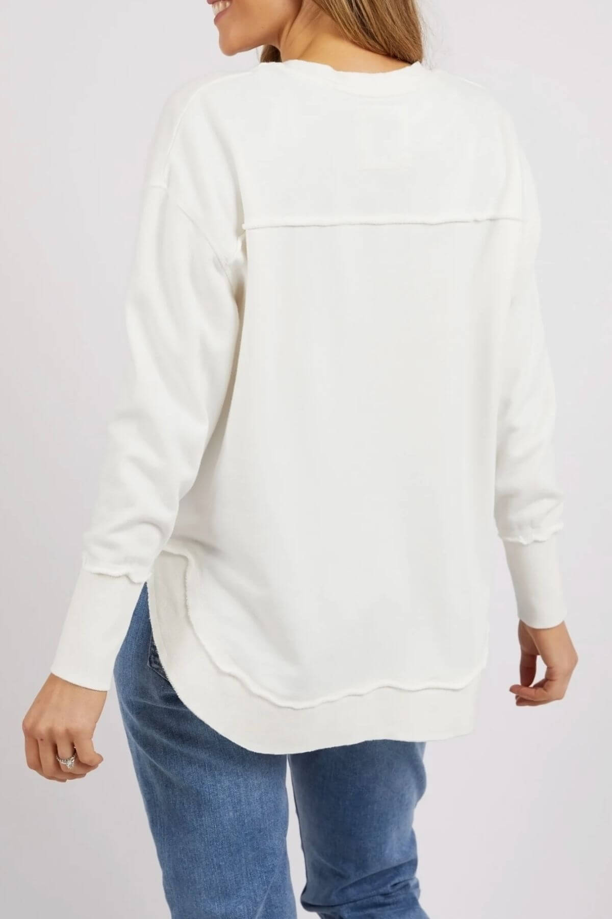 A woman with her hair down, smiling with her back to the camera, wears blue jeans and the Foxwood Delilah Crew—a long-sleeve, 100% cotton sweatshirt featuring a relaxed fit and curved hem. She has a ring on her right hand.
