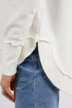 A person wears the Foxwood Delilah Crew, a white long-sleeve top with a raw curved hem, over light blue jeans. The focus is on the lower side of the shirt and top of the jeans, highlighting its 100% cotton comfort.