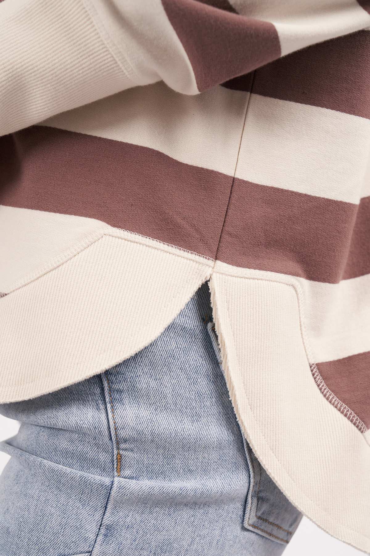 Close-up of a person wearing the Foxwood Delilah Stripe Crew, partially tucked into light blue jeans. The beige and brown striped sweater, made from 100% cotton, features a small side slit at the hem, adding a casual and relaxed look to the outfit.